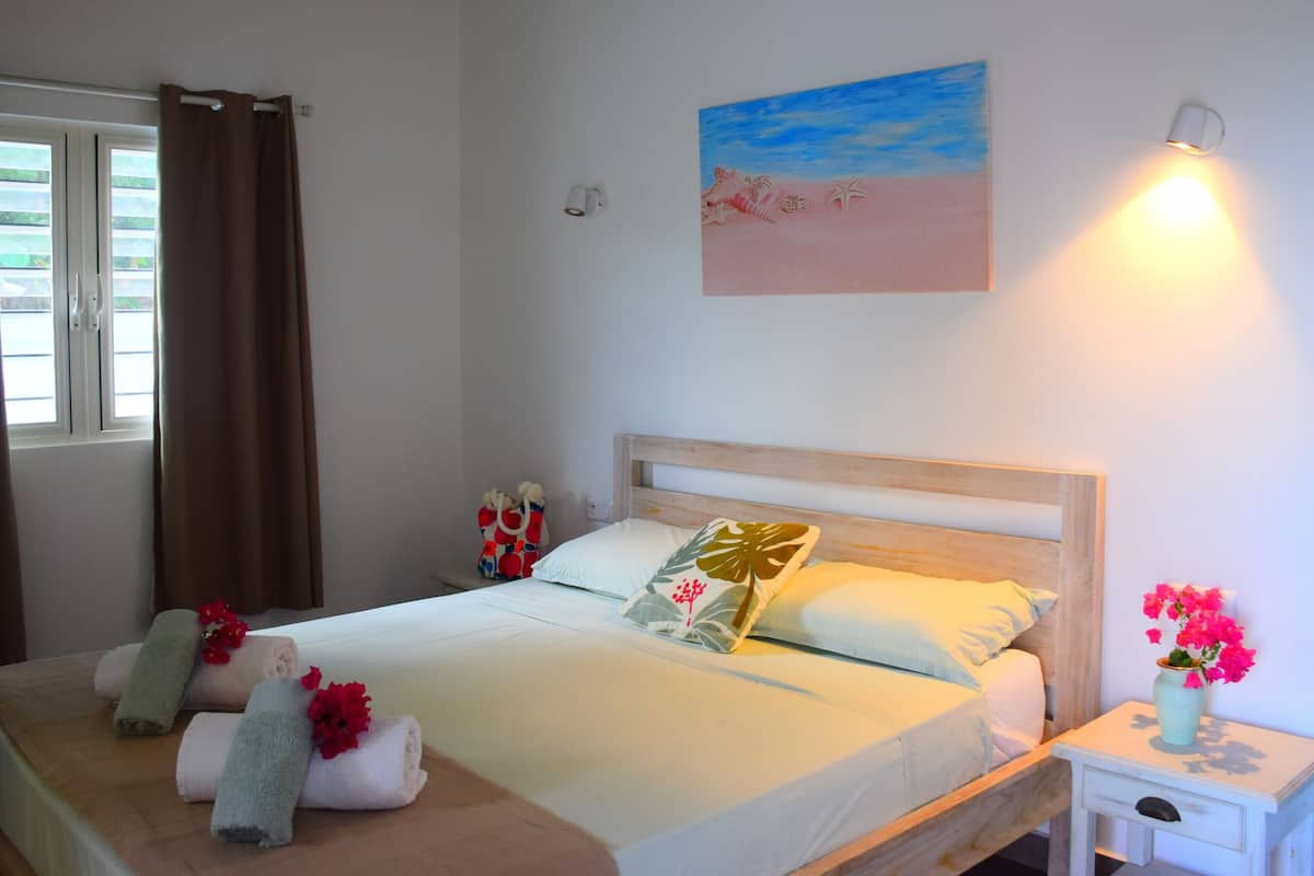 Ein ordentlich bezogenes Bett mit Handtüchern und Kopfkissen, ein Nachttisch mit Blumen in einer Vase, ein Bild an der Wand und ein Fenster mit Vorhängen.