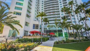 A modern hotel with multiple floors, outdoor seating areas with red umbrellas, and palm trees. - Apart Suites By Hilton. Top View. (Fort Lauderdale)
