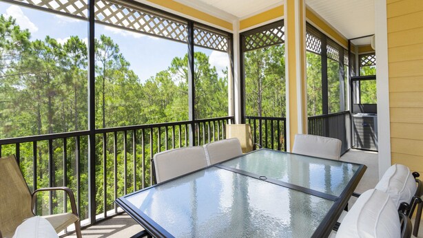 A covered outdoor patio with a glass table and chairs, surrounded by a black railing, overlooking a forested area. - 9.1 miles from Disney World, Sleeps 8-10, heated pool, hot tub, private balcony (Kissimmee)