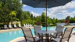 A poolside area with a table and chairs, surrounded by a pool, greenery, and a view of a lake. - 9.1 miles from Disney World, Sleeps 8-10, heated pool, hot tub, private balcony (Kissimmee)