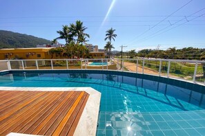 A swimming pool with a wooden deck, surrounded by a white fence, with a view of palm trees and buildings in the background. - BEAUTIFUL APARTMENT 26 - with sea view, two parking spaces, we have other options. (Ubatuba)