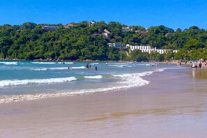 A beach with waves, people, and buildings on a hillside.