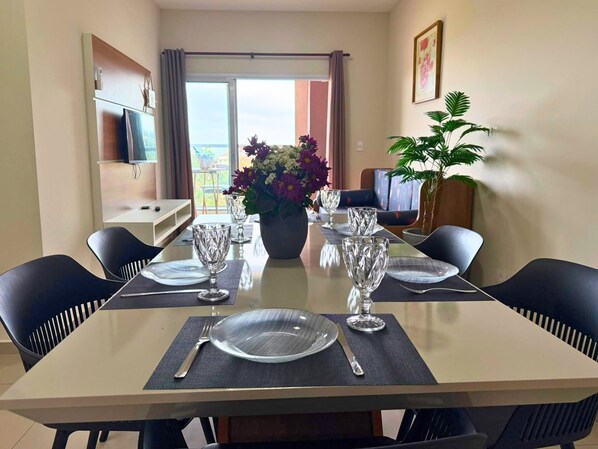 A dining area with a table set for four, including plates, cutlery, and glasses. There are chairs around the table, a potted plant, and a television on a shelf.