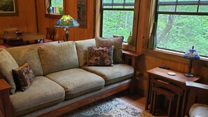 Un salón acogedor con un sofá de madera, cojines estampados, una lámpara de vitral y vistas al bosque a través de amplias ventanas.