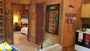 Una habitación acogedora con paredes de madera, una cama con cabecera azul, una mesita con una lámpara y una kitchenette con estufa y toallas colgadas.