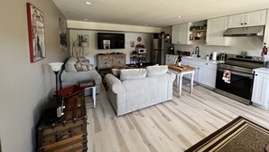 Un salon moderne avec un canapé gris, une table basse et une radio ancienne. La cuisine est équipée de meubles blancs, d’un réfrigérateur en acier inoxydable et d’une cuisinière.