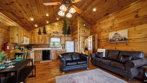 A log cabin interior with a dining area, two sofas, a kitchen, and a ceiling fan.
