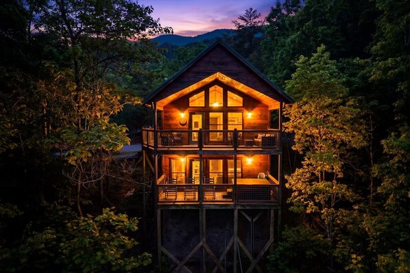 A two-story wooden cabin with a balcony, surrounded by dense forest.