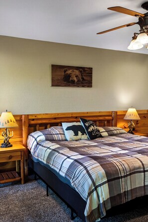 Une chambre à coucher avec une tête de lit en bois, une couvre-lit à carreaux, deux lampes de chevet, un ventilateur de plafond et une décoration murale sur le thème des ours.