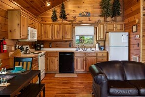A cabin interior with wooden walls, a kitchen with wooden cabinets, a dining area with a table and chairs, and a leather sofa.