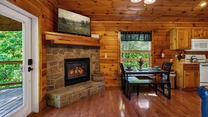 A wooden cabin interior with a stone fireplace, dining area, kitchen, and a television mounted above the fireplace.