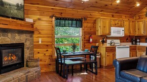 A cozy cabin interior with a stone fireplace, wooden walls, a dining area, and a kitchen with wooden cabinets and a microwave.