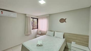 Quarto com cama, mesa de cabeceira, ar condicionado fixado na parede, janela com cortinas e um objeto decorativo na parede.