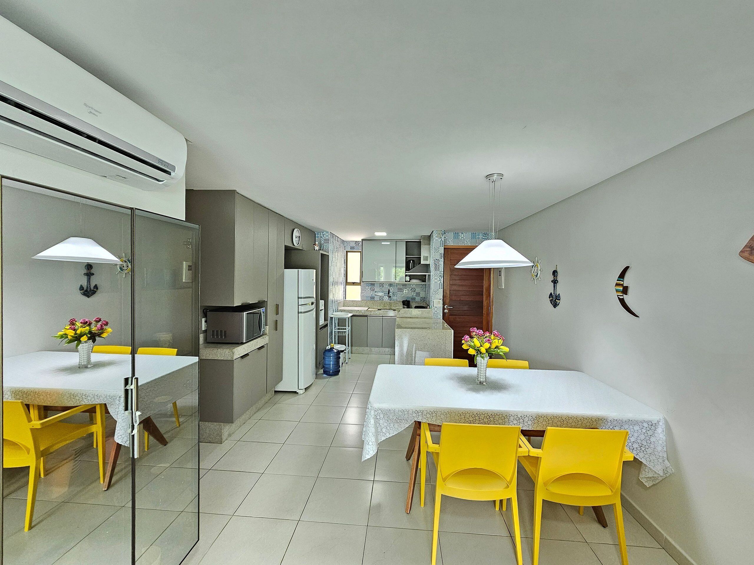 Een moderne keuken met een magnetron, koelkast en een eethoek met gele stoelen en een witte tafel.