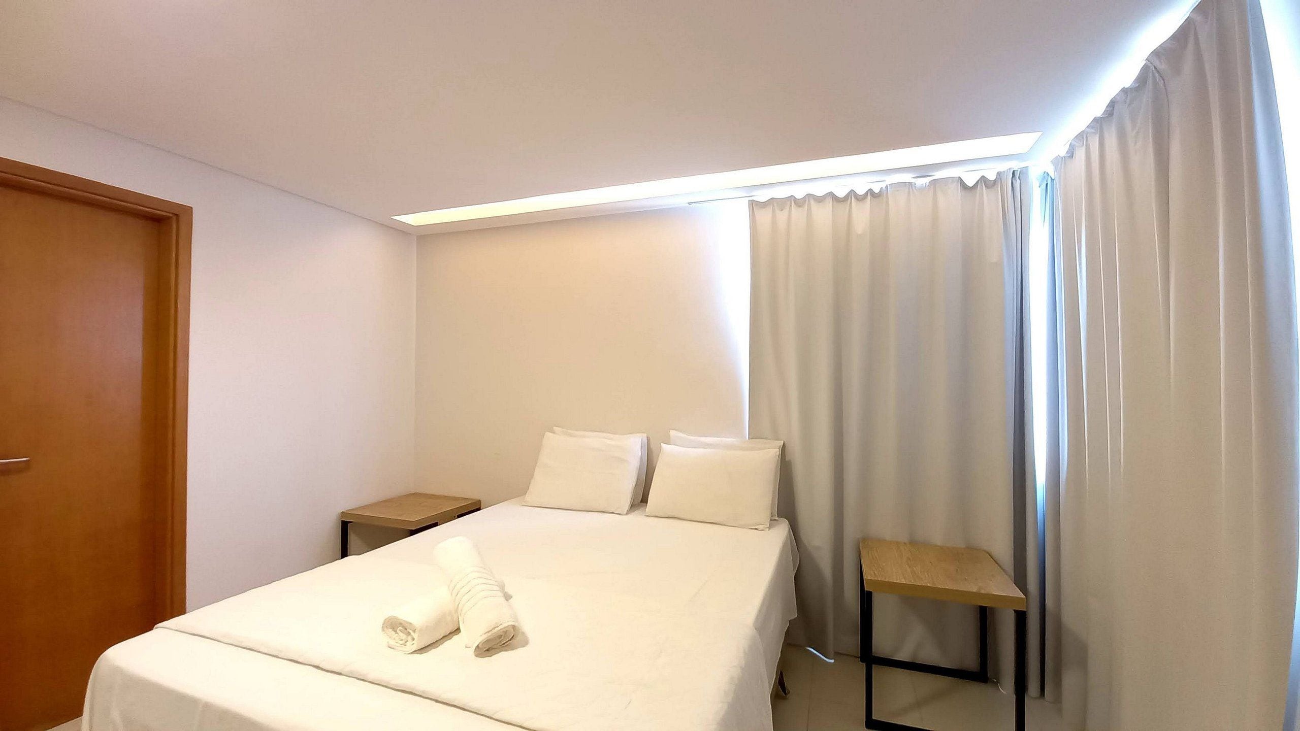 Habitación de hotel con una cama, dos almohadas, una mesita de noche de madera, una pequeña mesa y una puerta.