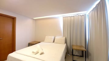 A hotel room with a bed, two pillows, a wooden nightstand, a small table, and a door.
