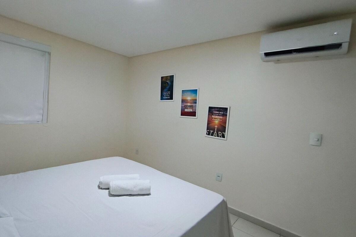 Una cama individual con sábanas blancas y dos toallas enrolladas, una ventana con persianas y un equipo de aire acondicionado en la pared.