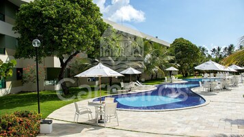 Zona de piscina con sillones de descanso y sombrillas, rodeada de follaje y un edificio.