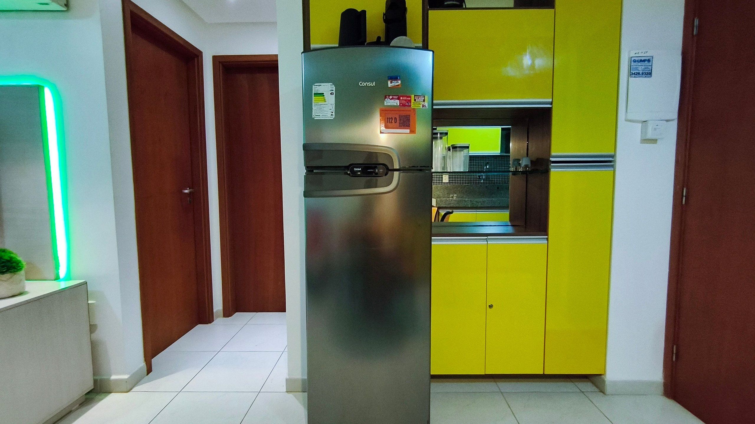 Cozinha com geladeira de aço inoxidável, armário amarelo e parede iluminada em verde.