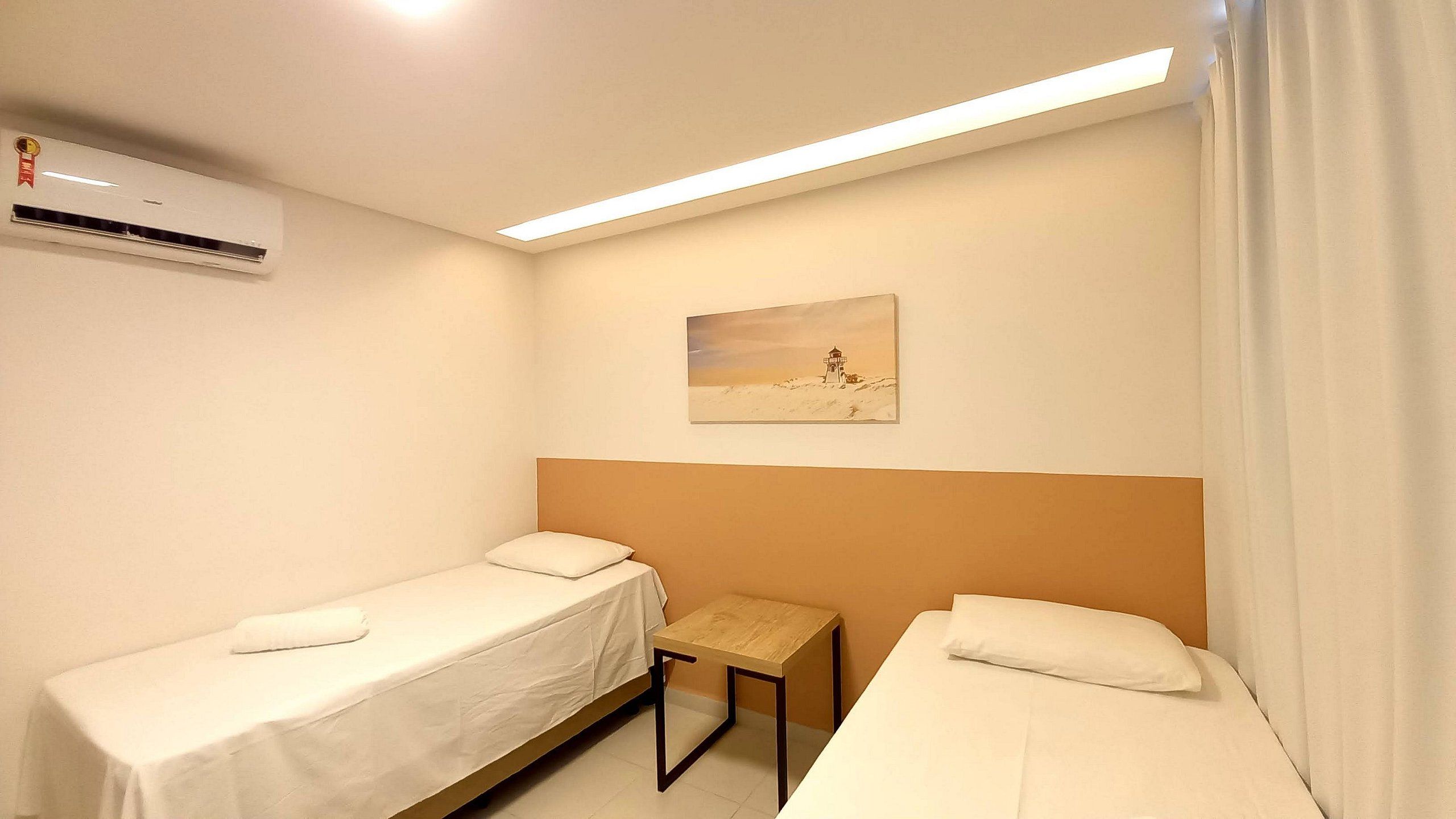 A hotel room with two single beds, a small table, an air conditioning unit, and a painting on the wall.