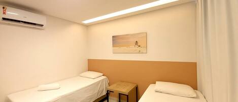 A hotel room with two single beds, a small table, an air conditioning unit, and a painting on the wall.