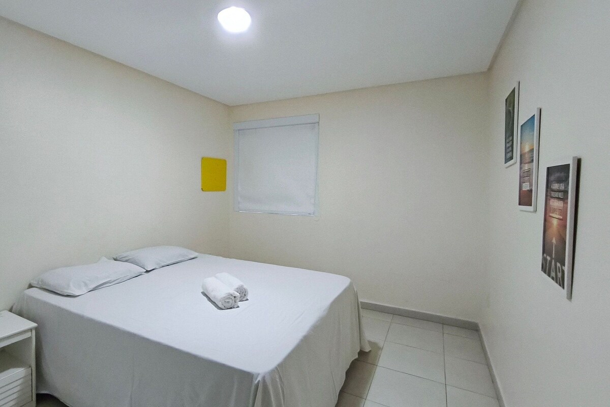 Una cama individual con sábanas blancas, una mesita de noche pequeña, una ventana con persianas y una pared con cuadros enmarcados.