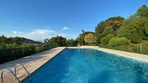 Una piscina rectangular amb un tram buidat de fusta i baranes de metall, envoltada per una tanca verda i vegetació abundant.