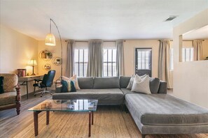 Relax in this inviting living space with a sectional sofa, coffee table, and desk, surrounded by large windows and wall-mounted shelves.