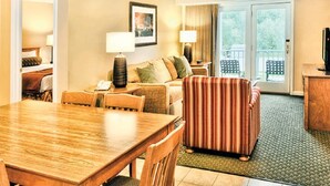 Habitación de hotel con una mesa de comedor, un sofá, un sillón a rayas, un escritorio pequeño con una lámpara y vistas al exterior a través de una puerta corrediza de vidrio.
