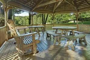 Situated on a wooden deck, this gazebo offers a serene seating area with a bench and two picnic tables surrounded by lush greenery.