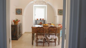 A dining area with a wooden table, two chairs, a chandelier, and a view through an arched doorway to another room.