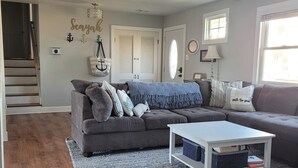 Sala de estar con un sofá gris, una mesa de centro blanca y un sillón de cuero negro. Se ve una escalera que sube a otra habitación.