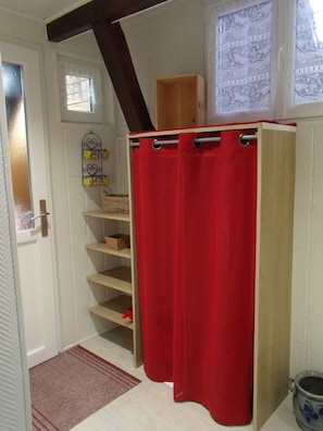 Un baño con cortina de ducha roja, repisas de madera, un ventanal pequeño con cortinas de encaje y una puerta con manija de metal.