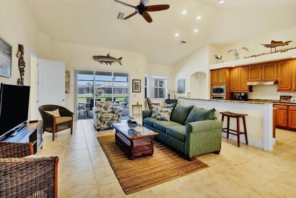 A spacious living room with a large sofa, a coffee table, and a television. There is a kitchen area with wooden cabinets and a bar stool.