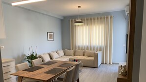 A modern living room with a wooden dining table, beige sofa, and light-colored curtains.