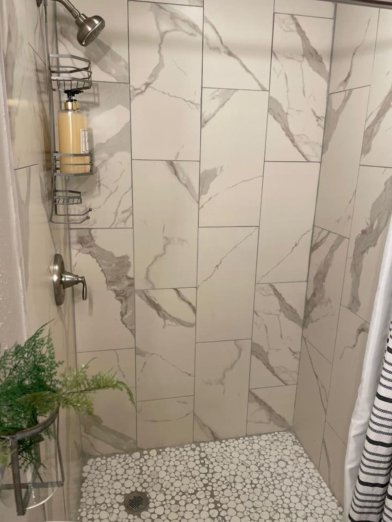A shower with marble tiles, a glass shelf with toiletries, and a pebble-patterned floor.