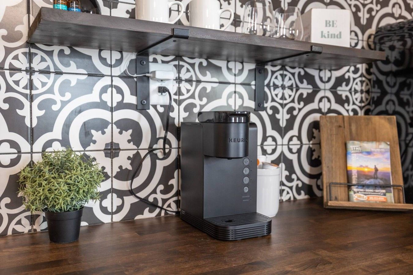 Een koffiezetapparaat op een aanrecht met een decoratieve spatwand en een kleine potplant.