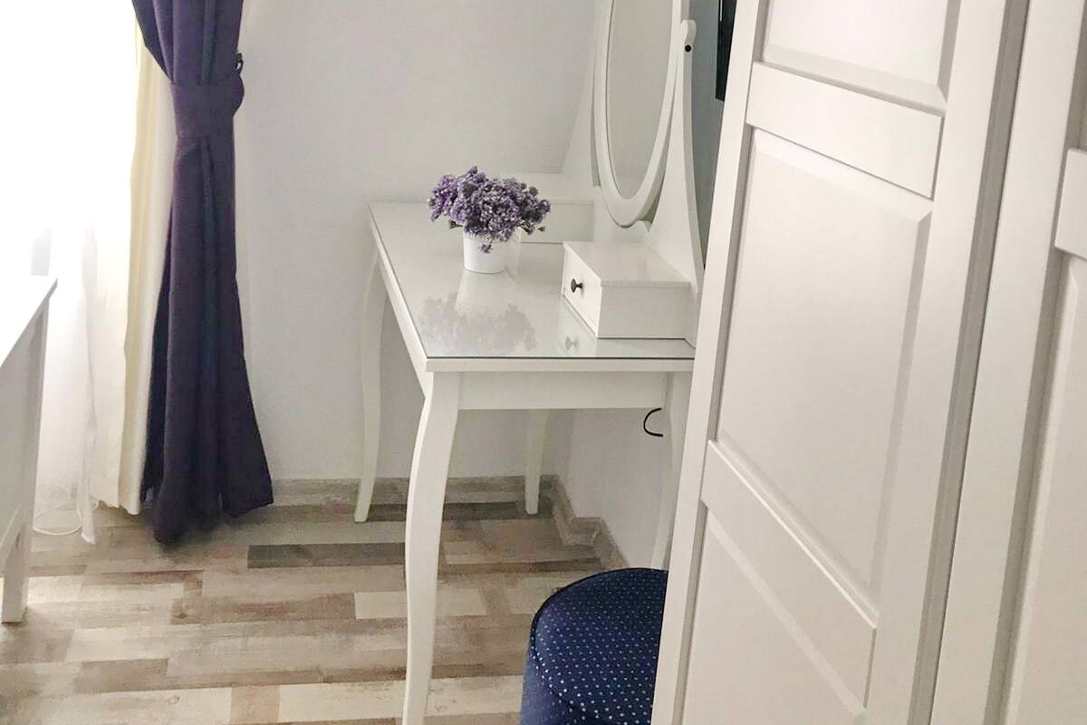 A small, white vanity with a mirror and a vase of flowers, a blue chair, and a wooden floor.