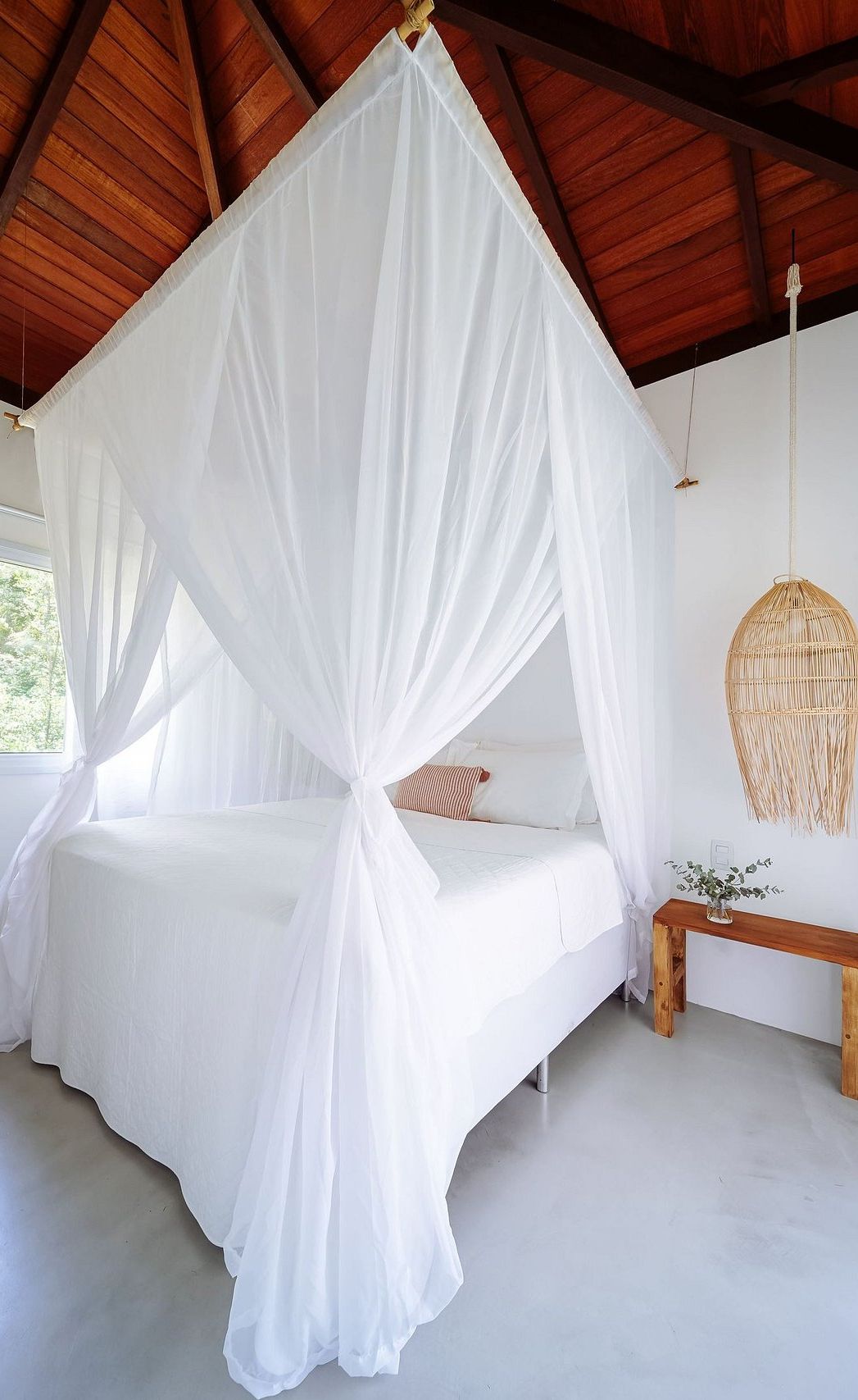 Ein Zimmer mit einem Bett unter einem weißen Baldachin, einem hängenden Stuhl aus Rattan und einer kleinen Holzbank.