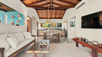 A spacious living area with a wooden ceiling, a sofa, a dining table, and a kitchen in the background.