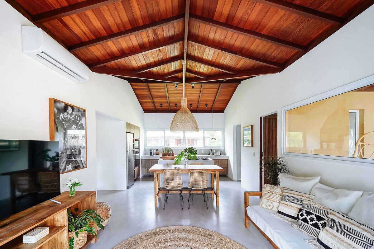 A spacious living area with a wooden ceiling, a dining table with chairs, a sofa, and a TV on a wooden stand.