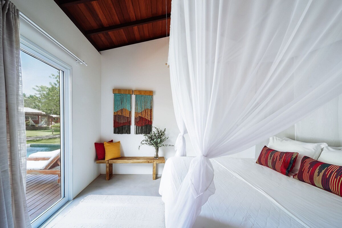 Quarto com cama de dossel, um banco de madeira, um banco com almofadas e vista para uma piscina lá fora.
