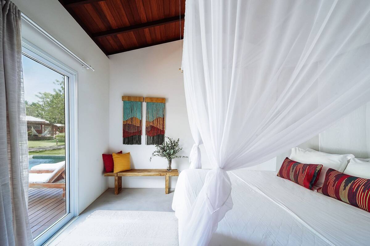 A bedroom with a canopy bed, a wooden bench, a bench with cushions, and a view of a pool outside.