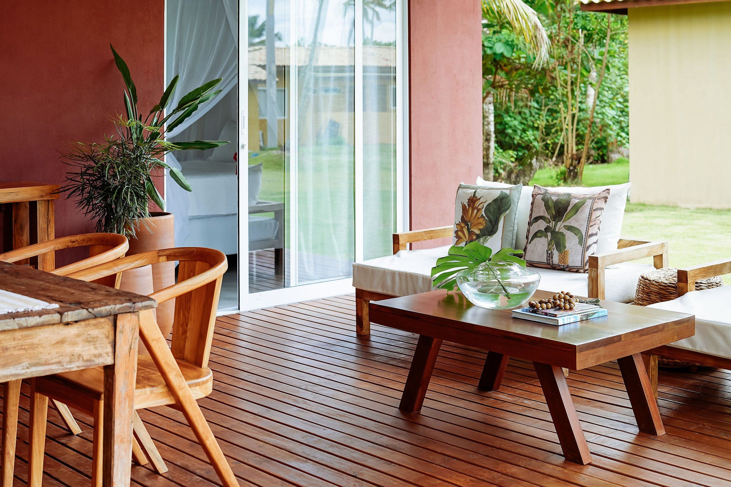 A wooden deck with a sofa and chairs, a table with a plant, and a view of greenery through a glass door.