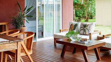 A wooden deck with a sofa and chairs, a table with a plant, and a view of greenery through a glass door.