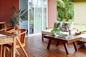Um deck de madeira com um sofá e cadeiras, uma mesa com uma planta e vista para o verde através de uma porta de vidro.