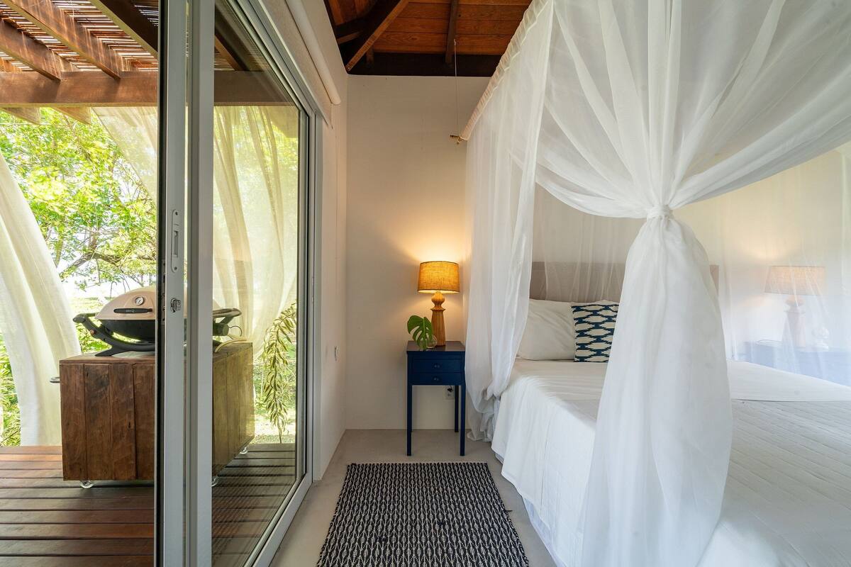A bedroom with a canopy bed, a bedside table with a lamp, and a view of greenery outside.