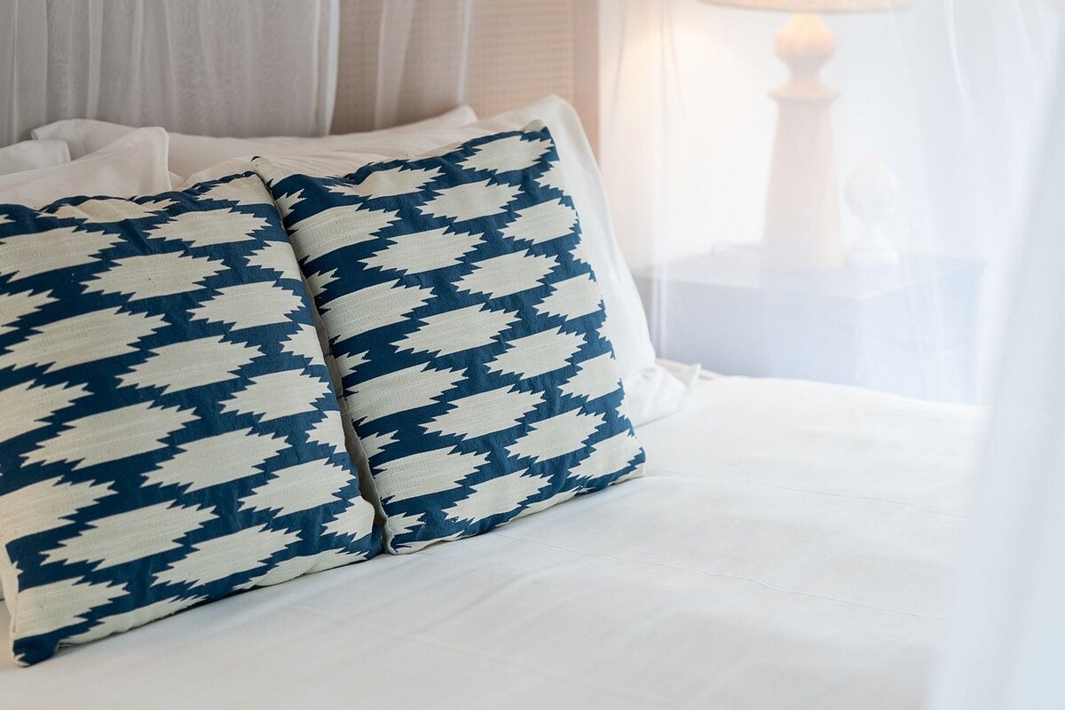 Cama com roupa de cama branca e almofada estampada em azul e branco.