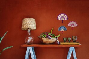 A wooden table with a woven lamp, a bowl of fruit, and decorative fans against a red wall. - Orixás Residence (Cairu)