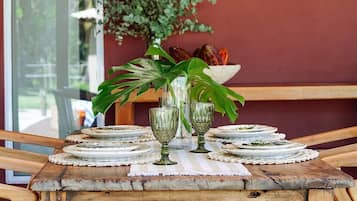A rustic wooden table with a woven pendant light, surrounded by green glassware and plates on a weathered tablecloth.